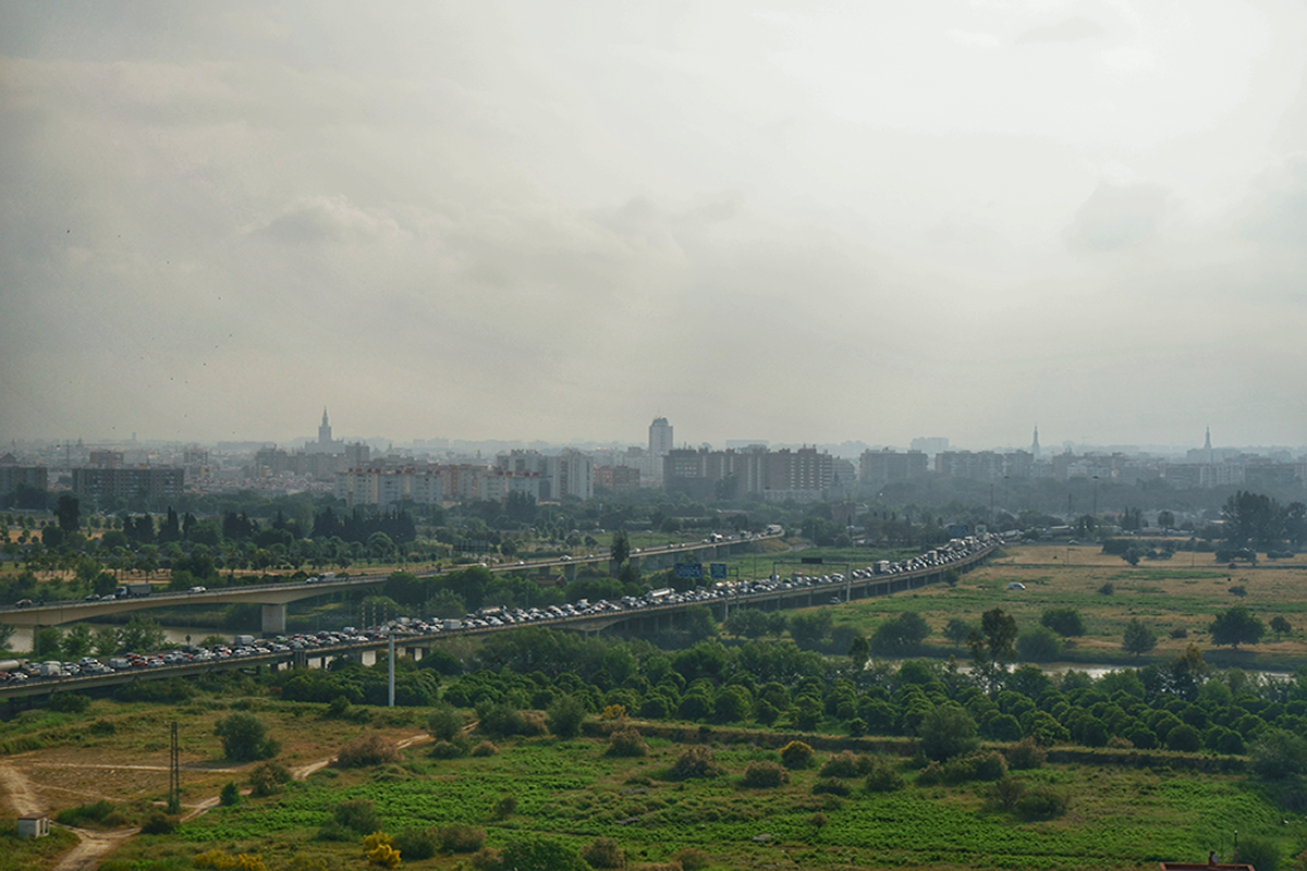  Movilidad residencia - trabajo en la aglomeraci&oacute;n urbana de Sevilla (los accesos a Sevilla desde el Aljarafe) 
Salvando ciertas distancias, esta fotograf&iacute;a es heredera de las muchas vistas panor&aacute;micas que desde el siglo XVI representaban la ciudad de Sevilla desde el Aljarafe. En casi todas ellas el r&iacute;o aparec&iacute;a simb&oacute;licamente bien representado, como eje y motor econ&oacute;mico de la ciudad moderna. Una de las principales novedades que incorpora esta vista contempor&aacute;nea &mdash;m&aacute;s all&aacute; de un skyline muy modificado donde no ha perdido protagonismo la Giralda&mdash; centra el tema de la fotograf&iacute;a: se trata de los dos viaductos sobre el nuevo cauce del r&iacute;o Guadalquivir, que simbolizan la victoria de la ingenier&iacute;a civil sobre los elementos limitantes del medio natural. Sin embargo, una interpretaci&oacute;n m&aacute;s retorcida de esta panor&aacute;mica nos lleva a pensar que ese mismo progreso t&eacute;cnicocient&iacute;fico que ha permitido conectar la ciudad central con su corona periurbana salvando el r&iacute;o, ha originado nuevos problemas de gesti&oacute;n territorial debido al intenso tr&aacute;fico que soportan estas carreteras. Los viaductos, que evitan levitando la llanura inundable, &ldquo;ahogan&rdquo; al ciudadano-conductor que intenta alcanzar Sevilla cada ma&ntilde;ana (v&eacute;ase la anterior fotograf&iacute;a). Ir en contra de la Naturaleza vuelve a ser aqu&iacute; una lecci&oacute;n no del todo bien aprendida...